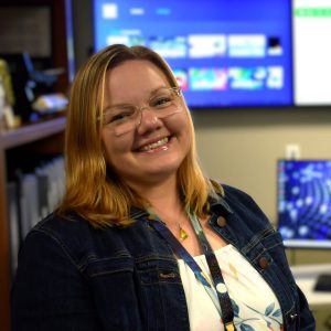Smiling woman with blonde hair and glasses wearing a denim jacket, standing in front of large screens in a tech or office setting.
